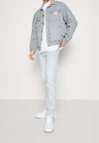 Person med en randig denimjacka, vit t-shirt och ljusblå jeans står mot en enfärgad bakgrund med händerna i fickorna.