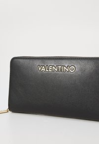 Juodos odinės piniginės, turinčios lygią tekstūrą, su auksiniu "VALENTINO" logotipu ir užtrauktuku. Stačiakampio formos su auksinio metalo akcentu.