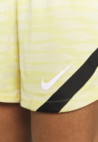 Pantalones cortos amarillos con un patrón ondulado, hechos de una tela ligera, con un acento negro y un logo de Nike blanco en el lado inferior.