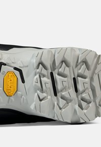 Svart skosåle med dype spor, med et Vibram-logo i gult. Teksturert grå gummi med vinklete mønstre og svarte detaljer.