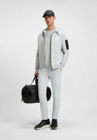 Felpa grigia chiara con zip e dettagli neri, abbinata a pantaloni grigi chiari e sneakers nere. Borsa a tracolla nera in mano. Design semplice e sportivo.