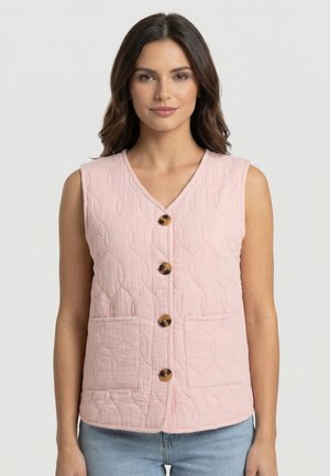 Donna che indossa un gilet trapuntato senza maniche di colore rosa chiaro con quattro bottoni in tartaruga e due tasche frontali, abbinato a jeans azzurri.