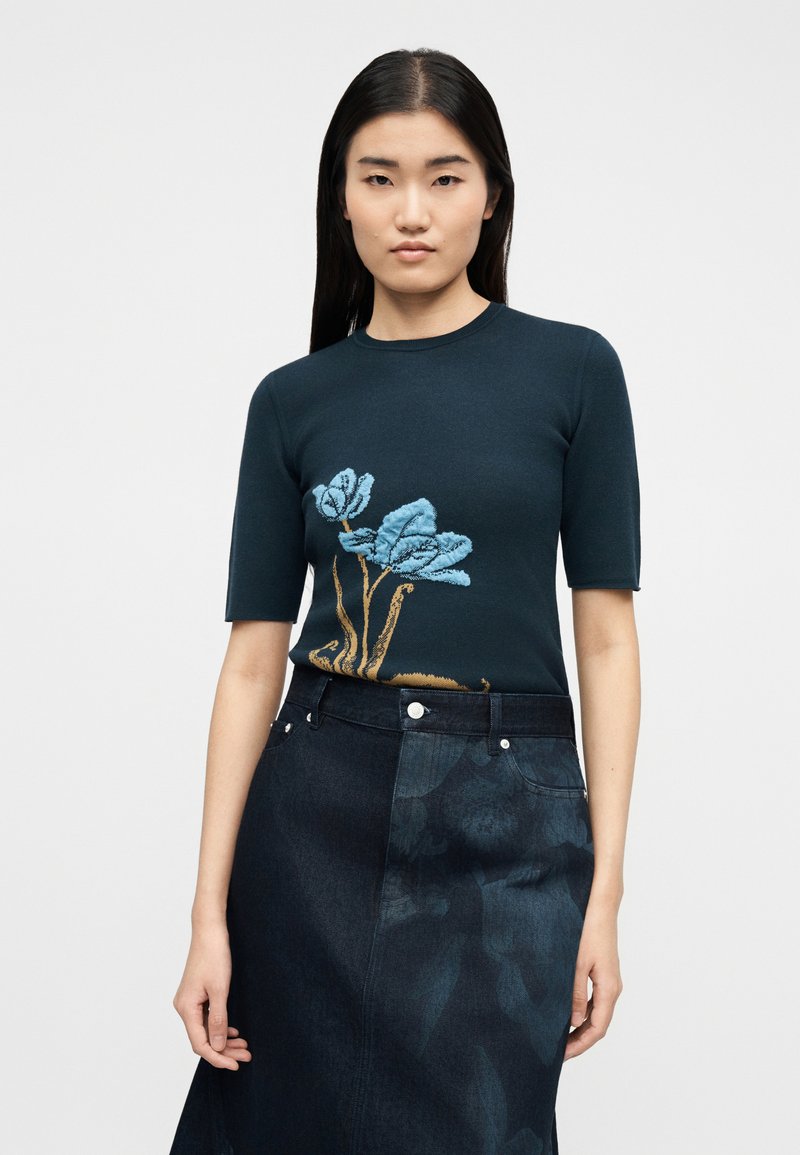 Jonge vrouw met lang donker haar draagt een marineblauw overhemd met korte mouwen en een blauw bloemmotief en een donker denim rokje met een subtiele bloemenprint.