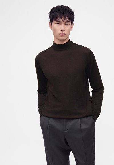 Jeune homme aux cheveux courts noirs portant un pull à col roulé marron foncé et un pantalon plissé gris foncé, debout les mains dans les poches.