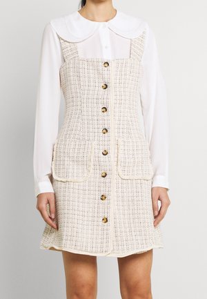 Frau trägt ein ärmelloses, beige-schwarz kariertes Hemdkleid mit Knöpfen und Taschen vorne über einer weißen Langarmbluse mit rundem Kragen.