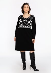 Une femme présente une robe noire à manches longues ornée de broderies florales blanches, associée à des bottes à talons noires montantes, sur un fond uni.