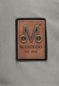Parcha de cuero marrón con un borde de fieltro negro que muestra un logo en relieve con una "M" y el texto "MARIKOO EST 2016", sobre un fondo de tela gris.