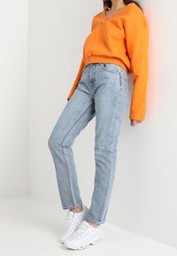 Sweatshirt orange surdimensionné, jeans taille haute bleu clair et baskets blanches. Le tissu est lisse ; les jeans ont une coupe ajustée et des détails subtils.