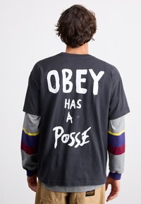 Tricou din bumbac negru, cu text alb care spune "OBEY ARE O POSADA" pe spate. Mâneci lungi în gri cu dungi roșii, galbene și albastre la manșete.
