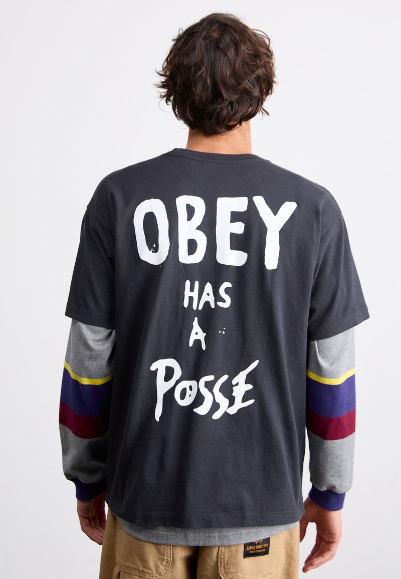 Tricou din bumbac negru, cu text alb care spune "OBEY ARE O POSADA" pe spate. Mâneci lungi în gri cu dungi roșii, galbene și albastre la manșete.