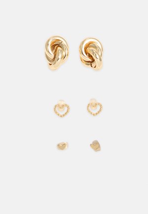 Trois paires de boucles d'oreilles clous en or disposées verticalement : paire du haut design nœud, paire du milieu forme cœur ouverte, paire du bas forme cœur pleine.