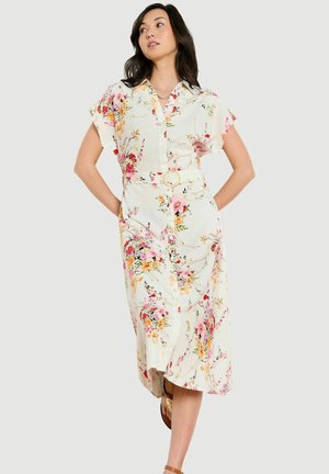 Noor naine kannab valget lühikeste varrukatega lillelist midi-kleit koos sobiva vööga, seisab käed selja taga.