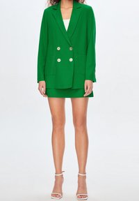 Tuuli-painettu vihreä kaksirivinen bleiseri, jossa on kaksi etutaskua ja hopeiset napit, yhdistettynä sopivaan lyhyeen hameeseen ja valkoisiin korkokenkiin.