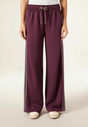 Mujer con pantalones deportivos sueltos en color granate con rayas laterales beige, cintura ajustable con cordón, camiseta blanca y zapatos beige tipo slip-on, de pie.