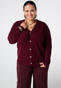 Bordeaux cardigan met hartvormige knopen, v-hals ontwerp, zachte textuur, gedragen over bijpassende bordeauxbroek. Accenten van goudkleurige sieraden.