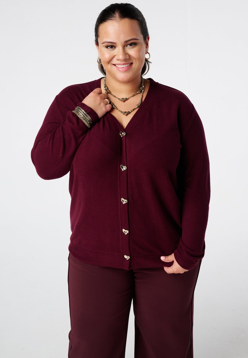 Bordeaux cardigan met hartvormige knopen, v-hals ontwerp, zachte textuur, gedragen over bijpassende bordeauxbroek. Accenten van goudkleurige sieraden.