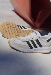Weißer Adidas Sneaker mit schwarzen Streifen, beiger Sohle und Schnürsenkeln, neben einem umgedrehten Schuh, der seine strukturierte Außensohle zeigt.