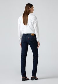 Jeans in denim blu scuro con un taglio slim, caratterizzati da tasche posteriori, una leggera scoloritura e un'etichetta in pelle marrone sulla vita. Abbinati a una camicia bianca.