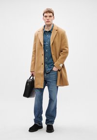 Μπεζ παλτό από μαλλί με κομμένες πέτες, τζιν πουκάμισο, μπλε τζιν και μαύρα σουέτ παπούτσια. Κρατώντας μια μαύρη δερμάτινη τσάντα. Απλός, δομημένος σχεδιασμός.