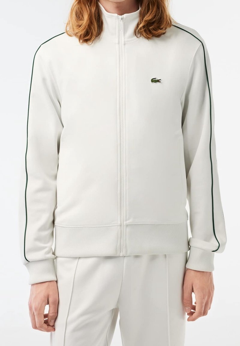 Giacca bianca con zip, rifinita con piping verde, polsini e orlo a costine. Presenta un piccolo logo verde di Lacoste sul lato sinistro del petto. Texture liscia.