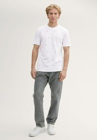 Camiseta blanca con un patrón geométrico de pequeños triángulos de colores, combinada con jeans gris claro y zapatillas blancas. Ajuste y estilo casual.