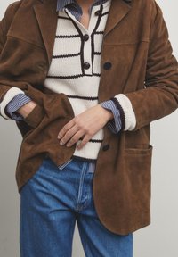 Giacca in suede marrone con colletto a punta sopra un top in maglia bianca con linee marroni. Abbinata a jeans in denim blu, con dettagli testurizzati.