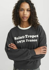Tmavě šedá mikina s krémovým límcem. Na přední straně je výrazný bílý text "Saint-Tropez 1970 Francie." Volný střih, žebrované manžety a měkká textura.