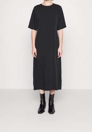 Personne portant une robe noire mi-longue à manches courtes et des bottines à talons noires, debout devant un fond blanc uni.