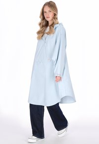 Világoskék vízálló kabát kapucnival, elülső zsebbel és gombos részletekkel. Laza szabás, hosszú ujjakkal és ívelt aljjal. Sötét farmernadrág fölött viselve.