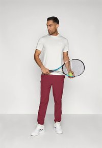 Męski strój sportowy: jasnoszara koszulka z krótkim rękawem w wzory oraz bordowe spodnie. Trzyma rakietę tenisową i piłkę, ma na sobie białe tenisówki.