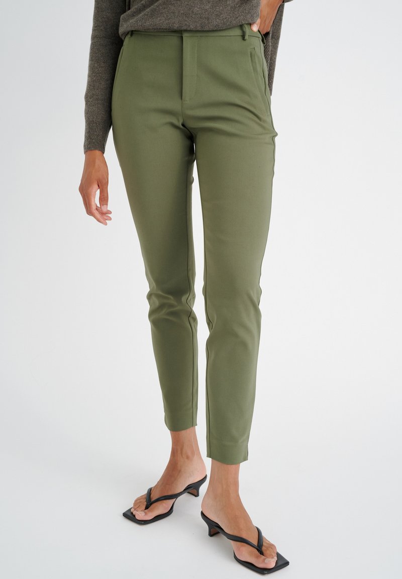 Pantaloni eleganti verde oliva con vestibilità slim, texture liscia, tasche laterali e vita media. Abbinati a ciabatte con tacco nere.