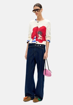 Kobieta w za dużych ciemnoniebieskich dżinsach, kremowym kardiganie z czerwonym wzorem, w paskowanej koszuli, okularach przeciwsłonecznych, brązowych butach, trzymająca małą różową torebkę.