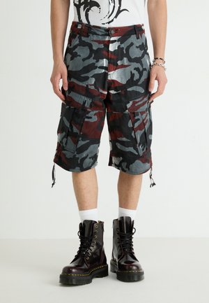 Camo Cargo-Shorts in Grau, Rot und Schwarz, mit mehreren Taschen und Kordelzugsäumen, kombiniert mit schwarzen Schnürstiefeln.