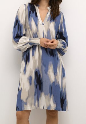 Femme portant une robe ample à manches longues et col en V avec des motifs abstraits bleu, beige et noir, debout devant un fond blanc uni.