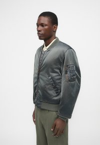 Casaco bombardeiro cinza feito de material semelhante ao cetim, com fecho de correr na frente, punhos canelados e um bolso com fecho de correr na manga esquerda.