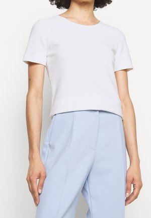 Kobieta ubrana w gładką białą bluzkę z krótkim rękawem, włożoną do wysokich, jasnoniebieskich, dopasowanych spodni.