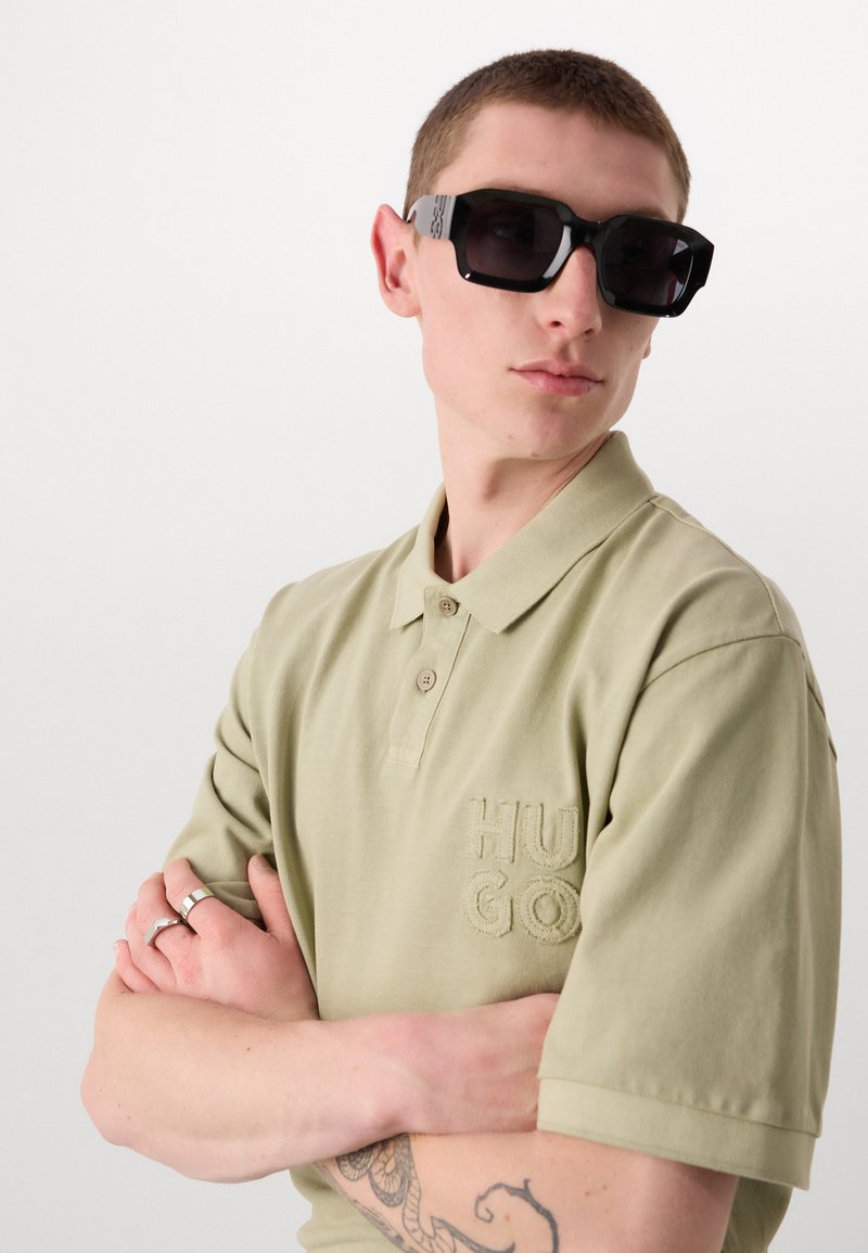 Polo shirt vert avec un logo texturé et deux boutons, associé à des lunettes de soleil géométriques noires. Le mannequin a les cheveux courts et des tatouages visibles.