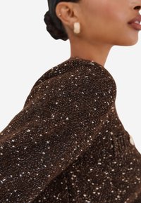 Profilo di una persona che indossa orecchini a cerchio dorati e un maglione di lana texture a macchie marrone scuro.