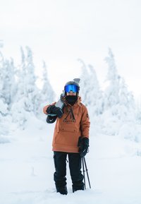 Orange snow jacket with a logo, black pants, gloves, and a black helmet. Skis on shoulder, snowy landscape in the background.