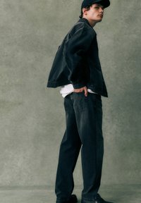 Dunkelblaue Jeansjacke und Hose in lockerem Schnitt, darüber ein weißes Hemd getragen, ergänzt durch eine schwarze Mütze. Texturiertes Material mit minimalen Details.