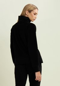 Chaqueta de terciopelo negro con cuello alto, mangas largas y un corte relajado. Presenta una textura suave y un diseño sólido sin patrones.