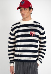 Hombre con gorra roja y jersey de punto a rayas azul marino y blanco con logo rojo "NY", combinado con pantalones gris con rayas finas, manos en los bolsillos.