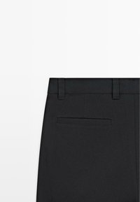 Pantalon formel noir avec passants pour ceinture et une unique poche passepoilée à l’arrière, visible de la partie haute arrière.