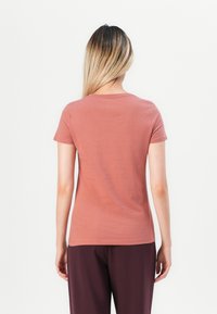 Woman with long blonde hair stands facing away, wearing a dusty rose short-sleeve shirt and dark purple pants against a white background.