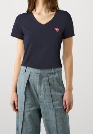 Frau trägt ein marineblaues T-Shirt mit V-Ausschnitt und kurzen Ärmeln mit rotem Guess-Logo auf der Brust sowie hoch taillierte graue plissierte Hose.