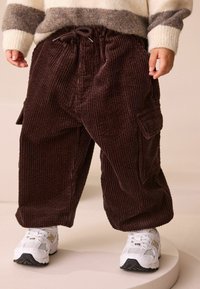 Bruna kadodräkter med avslappnad passform, med sidofickor och midjeband med elastisk dragsko, tillsammans med vita sneakers.