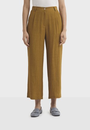 Donna che indossa pantaloni a gamba larga corta a vita alta color senape con bottoni frontali e mocassini grigi slip-on su sfondo bianco.