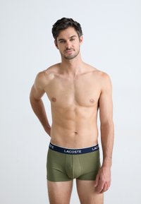 Groene stretch katoenen boxershorts met een marineblauwe tailleband waarop het Lacoste-logo staat, gemodelleerd door een man op een effen achtergrond.