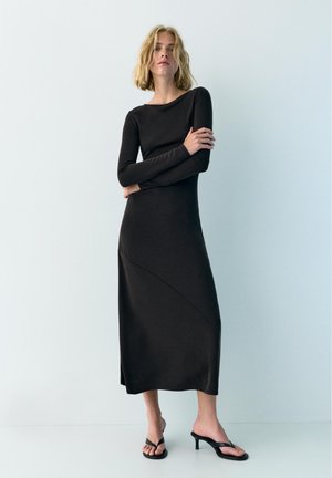 Vrouw met blond haar draagt een lange zwarte jurk met lange mouwen en zwarte open-toe sandalen met hakken, staand tegen een effen lichte achtergrond.