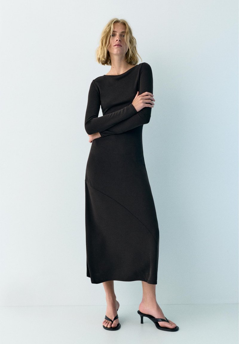 Femme aux cheveux blonds portant une longue robe noire à manches longues et des sandales à talons noires à bout ouvert, debout devant un fond clair uni.
